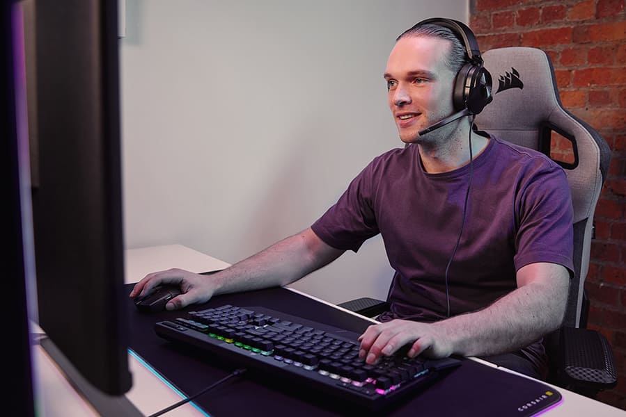 Persona sentada frente a un monitor de PC con auriculares para juegos con cable HS55 STEREO y un teclado para juegos K70 RGB PRO. 