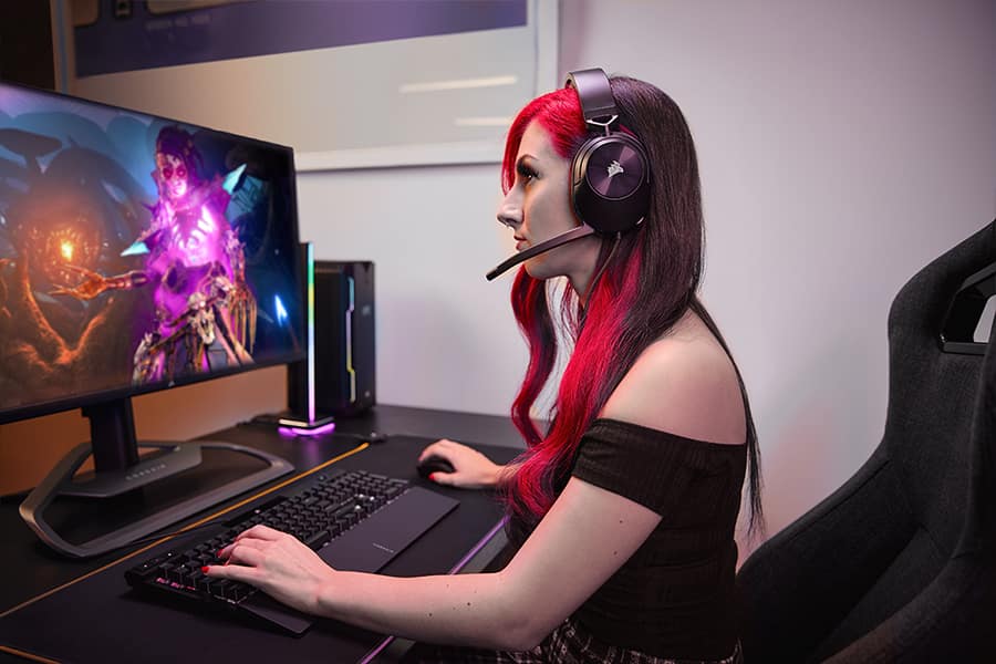 Một người phụ nữ đang chơi trò chơi điện tử trên PC chơi game đeo một cặp tai nghe chơi game có dây HS55 SURROUND màu đen và tay cầm chuột và bàn phím chơi game CORSAIR màu đen.