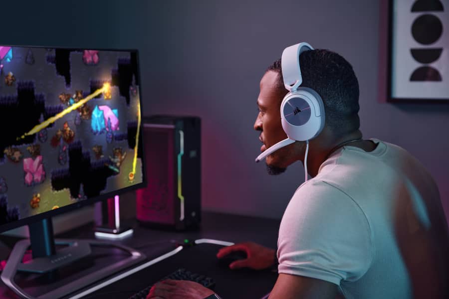 Một người đàn ông ngồi trước PC chơi trò chơi điện tử trong tai nghe chơi game HS55 SURROUND màu trắng. 