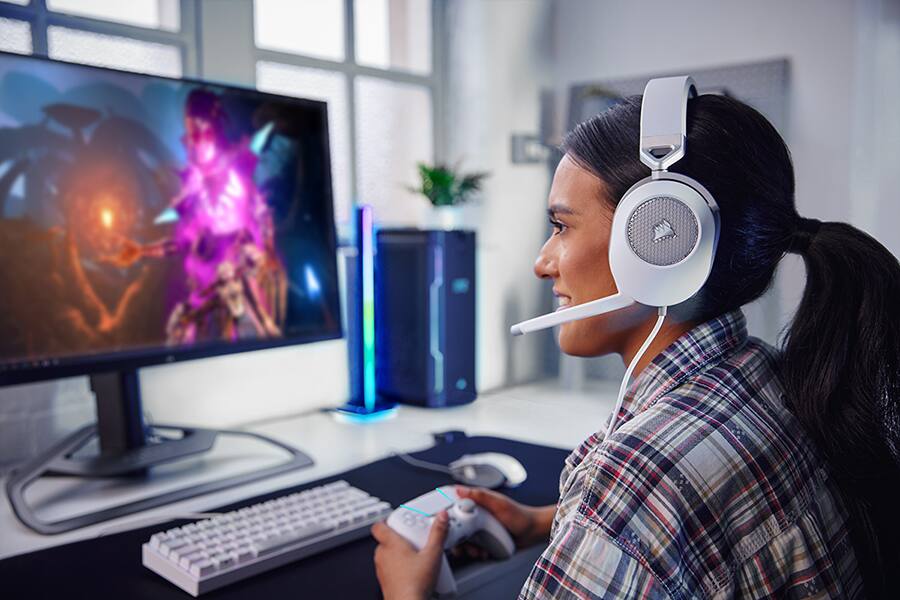 Mujer de perfil jugando a videojuegos con un mando frente a su PC para juegos y con auriculares para juegos con cable HS65 SURROUND blancos y un teclado y ratón blancos a juego. 