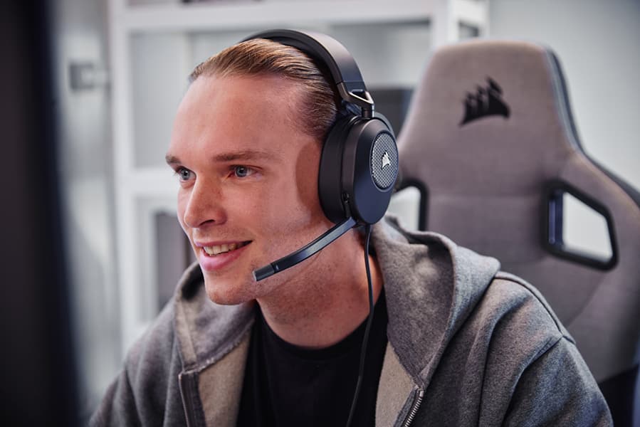 Hombre sonriendo sentado en una silla para juegos y mirando un monitor mientras lleva unos auriculares para juegos CORSAIR HS65 SURROUND de color negro. 