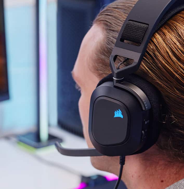 Người đàn ông đeo tai nghe chơi game có dây HS80 RGB USB trong khi chơi trò chơi điện tử với hệ thống đèn RGB trên biểu tượng cánh buồm Corsair.